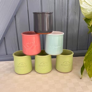 May include: A stack of six ceramic yogurt pots in various colours including dark grey, coral, light blue, and three light green pots. The pots feature embossed text reading La Fermiere and a heart design. The pots are arranged in a pyramid formation.