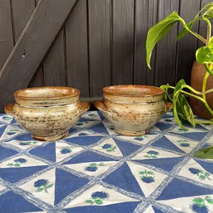 Op de afbeelding: Twee bruine keramische kommen met een gespikkeld glazuur. De kommen hebben een afgeronde vorm en twee handvatten. De kommen staan op een blauw-wit geblokt tafelkleed.