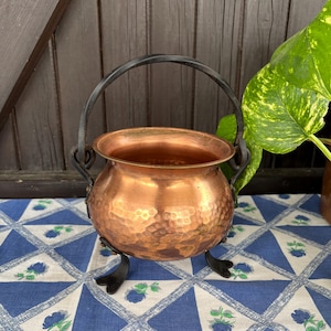 Puede incluir: Un pequeño caldero de cobre martillado con un asa y patas de hierro forjado negro. El caldero tiene un cuerpo redondo y un borde ancho. El fondo incluye un mantel con estampado azul y blanco y una puerta de madera.