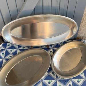 Könnte beinhalten: Drei ovale Servierplatten in Silberoptik. Die größte Platte befindet sich oben, darunter zwei kleinere. Jede Platte hat einen geriffelten Rand. Sie sind auf einer blau-weiß gemusterten Tischdecke angeordnet.