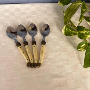 May include: Four stainless steel spoons with ivory-colored handles and brass accents. The spoons are arranged in a fan shape on a white checkered surface. The spoons are a kitchen utensil.