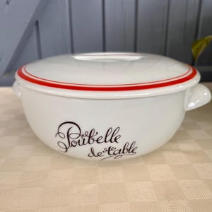 Op de afbeelding: Een witte keramische kom met bijpassend deksel, voorzien van een rode cirkelvormige streep rond de rand en twee handvatten aan de zijkanten. De voorzijde van de kom toont de zwarte cursieve tekst Poubelle de table, wat de functie als tafelafvalbak aangeeft.