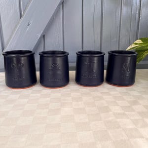 Peut inclure: Quatre tasses en céramique noir mat avec une base en terre cuite. Chaque tasse présente un symbole du zodiaque en relief et un mot correspondant : Bélier avec Belief, Balance avec Balance, Gémeaux avec Connection et Lion avec Lion.