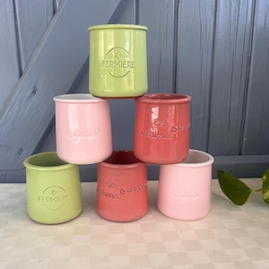 Puede incluir: Una pila de seis pequeños tarros cilíndricos de cerámica en tonos rosa, rojo y verde. Algunos tarros tienen texto en relieve, incluyendo "La Fermiere" y frases francesas. Los tarros están dispuestos en forma de pirámide.