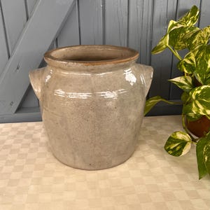 Op de afbeelding: Een vintage beige steengoed pot met twee kleine handvatten. De pot heeft een brede opening en een licht getextureerd oppervlak. De achtergrond heeft een lichtgrijze houten deur en een groen-gele plant.