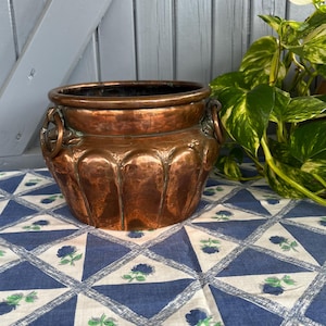 May include: A copper-coloured planter with a rounded body and two ring handles. The planter sits on a blue and white patterned tablecloth. Green and yellow leafy plant in the background. The planter is likely used for decorative purposes.
