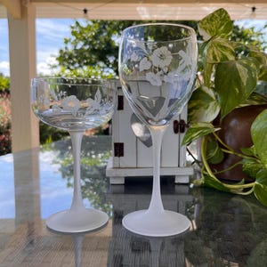 Könnte beinhalten: Zwei klare Weingläser mit geätzten Blumendesigns. Die Gläser haben mattierte weiße Stiele und Böden. Eines ist ein Standard-Weinglas, das andere ein kleineres Stielglas. Die Gläser stehen auf einem Glastisch, im Hintergrund ein weißes Dekorationselement.
