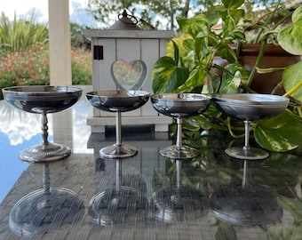 May include: Four silver champagne coupe glasses with a reflective surface. The glasses have a round bowl and a stem. The glasses are arranged in a row. The background includes a white decorative lantern and green plants.