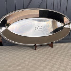 Könnte beinhalten: Ovaler, versilberter Servierteller mit Perlenrand. Das Tablett hat eine reflektierende Oberfläche und wird von einem Holzständer getragen. Das Tablett trägt in der Mitte den Text "Jean Couzon Orfevre".