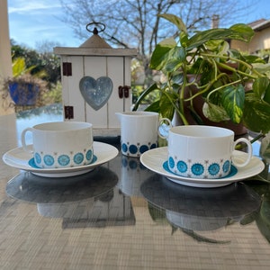 Op de afbeelding: Twee witte theekopjes met blauwe en witte bloemenpatronen op witte schoteltjes, een witte roomkan met een blauw en wit bloemenpatroon en een witte houten lantaarn met een hartvormig raam op een tafel met een geweven patroon.