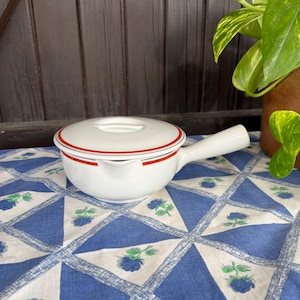 Op de afbeelding: Witte keramische sauspan met een rode en witte rand rond de rand en de handgreep. De sauspan heeft een deksel met een kleine knop bovenop.