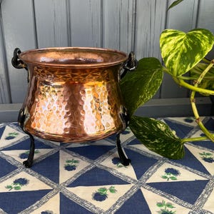 May include: A copper cauldron with a hammered finish, black iron handles and legs. The pot has a wide opening and sits on a blue and white floral tablecloth. Green and yellow leaves are visible in the background.