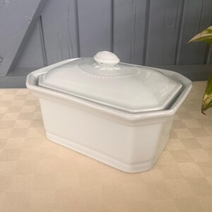 May include: A rectangular white ceramic butter dish with a matching lid. The lid features a small, rounded handle at the center and a decorative beaded border detail around the top edge. The base has smooth, straight sides and a clean finish.