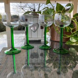 Op de afbeelding: Vier heldere glazen wijnglazen met groene stelen en voetjes. De glazen zijn in een rij op een tafel geplaatst, met een reflectie van de glazen zichtbaar.
