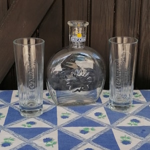 May include: A clear glass liquor bottle and two matching glasses with the brand name "RICARD" visible. The bottle is round with a flat base. The glasses are tall and cylindrical. The items are on a blue and white patterned tablecloth.