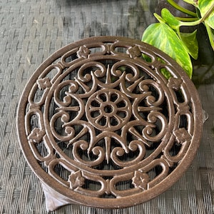 Peut inclure: Un dessous de plat rond en fonte brune avec un motif complexe et orné. Le dessous de plat a un bord surélevé et un fond plat.