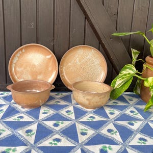 Op de afbeelding: Vier keramische kommen in tinten van bruin en wit met een gespikkeld glazuur. De kommen zijn gerangschikt op een blauw-wit gepatroneerd tafelkleed.