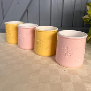 Może przedstawiać: Cztery małe ceramiczne kubki ustawione w rzędzie, naprzemiennie w kolorze żółtym i różowym. Każdy kubek ma białe wnętrze oraz wypukły, tłoczony wzór kwiatowy na zewnętrznej powierzchni, składający się z prostych kwiatów i małych listków.
