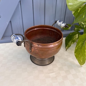 May include: A round copper bowl with a polished finish, featuring two curved metal handles attached to white ceramic grips decorated with blue floral patterns. The bowl rests on a circular pedestal base and has a smooth, open interior.