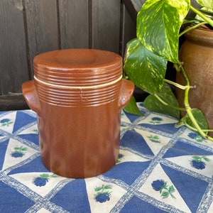 Può includere: Un vaso di ceramica marrone con un coperchio e due manici. Il vaso ha una striscia bianca attorno alla parte superiore e inferiore del coperchio. Il vaso è appoggiato su una tovaglia a quadri blu e bianchi.