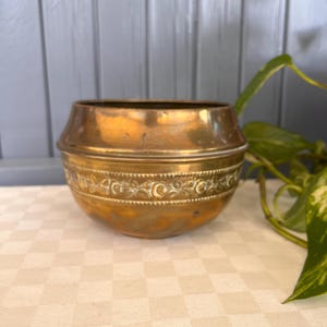 Puede incluir: Una maceta de latón con una banda floral decorativa. La maceta tiene una forma redondeada con una parte superior ligeramente ensanchada. El latón tiene una pátina cálida y envejecida, lo que sugiere que es una pieza antigua o vintage.