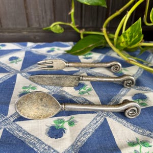 Pode incluir: Um conjunto de três talheres prateados com alças ornamentadas. Os talheres incluem um garfo, uma faca e uma colher. Os talheres estão dispostos sobre uma toalha de mesa estampada azul e branca.