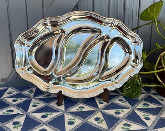 Pode incluir: Uma bandeja de servir prateada com uma borda ondulada e ornamentada e três compartimentos curvos. A bandeja é exibida em um suporte de madeira, colocada sobre uma toalha de mesa estampada em azul e branco. A bandeja reflete o ambiente.