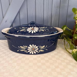 Könnte beinhalten: Eine marineblaue Keramik-Auflaufform mit Deckel. Die Form hat ein weißes Blumendesign mit orangefarbenen Zentren. Der Rand und der Griff sind weiß. Die Form ist oval und steht auf einer karierten Tischdecke.