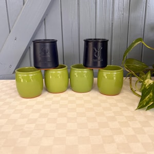 Peut inclure: Une collection de pots en céramique verts et noirs. Les pots verts sont disposés en rangée, tandis que les pots noirs sont empilés dessus. Les pots noirs présentent des symboles du zodiaque et du texte. Les pots sont sur une surface à carreaux.