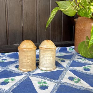 Könnte beinhalten: Ein Set aus zwei Keramik-Salz- und Pfefferstreuern mit brauner und weißer Glasur. Die Streuer stehen auf einer blau-weiß karierten Tischdecke mit Blumendesigns.