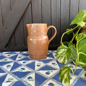 Puede incluir: Una jarra de cerámica marrón con una boca ancha y un asa. La jarra tiene una superficie texturizada y está sentada sobre un mantel azul y blanco.