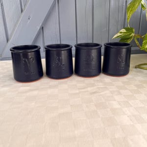 Op de afbeelding: Vier cilindervormige bekers van matzwart keramiek met een terracotta voet. Elke beker is voorzien van een in reliëf aangebracht sterrenbeeld en de bijbehorende naam: Aries, Leo, Balance en Leo. De bekers staan in een rij op een licht oppervlak.