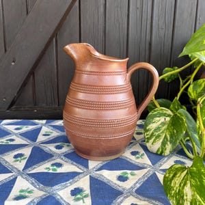 May include: A brown ceramic pitcher with a textured design. The pitcher has a wide mouth and a handle. It is sitting on a blue and white tablecloth.