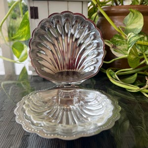 May include: A silver-coloured, hinged seashell-shaped trinket box. The box is open, revealing the interior. The shell design features detailed ridges and a scalloped edge. The box is likely made of metal and reflects light.
