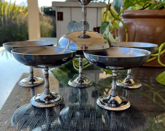 May include: A set of six silver-plated champagne coupes with a decorative design on the base. The coupes are arranged in a circle on a black surface.
