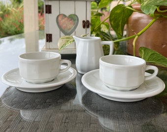 May include: Two white porcelain teacups and saucers with a white porcelain creamer. The cups and saucers have a simple, elegant design. The creamer has a small handle and a spout.