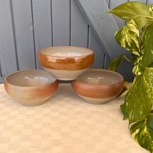 Peut inclure: Trois bols en céramique de tailles différentes. Les bols sont de couleur beige clair avec une bande orange-brun au milieu. Les bols sont disposés sur une nappe à carreaux, avec un fond en bois et une plante verte en arrière-plan.
