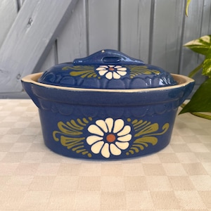 Puede incluir: Una cazuela de cerámica azul con tapa a juego, que presenta un motivo de margarita blanca pintado a mano con detalles de hojas verdes en el frente y la tapa. El recipiente tiene un borde festoneado y dos asas laterales integradas para sujetar.
