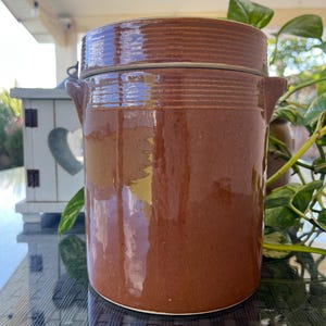 May include: A brown ceramic crock with a lid and two small handles. The crock has a glossy finish and horizontal ribbed detailing around the top. A metal wire secures the lid. The crock is cylindrical and sits on a white base.