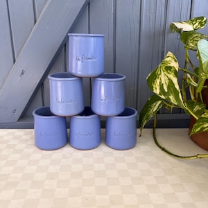 Könnte beinhalten: Ein Stapel aus sechs hellblauen Keramik-Joghurtbechern, die in einer Pyramide angeordnet sind. Jeder Becher trägt den geprägten Schriftzug la fermière auf der Vorderseite. Die Becher haben eine glatte, zylindrische Form mit einem leicht ausgestellten Rand.
