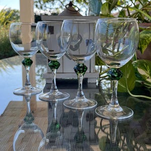 Puede incluir: Cuatro copas de vino transparentes con bases decorativas verdes están dispuestas sobre una superficie reflectante. Las copas tienen una forma de cuenco clásica y un tallo delgado. La base verde añade un toque de color al diseño elegante. El fondo está borroso.