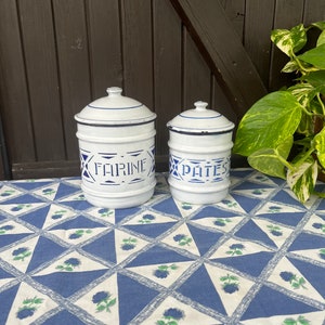 Può includere: Due barattoli vintage in smalto bianco con bordi e scritte blu. Il barattolo più grande dice "FARINE" e quello più piccolo dice "PATE". I barattoli sono su una tovaglia a motivi blu e bianchi.