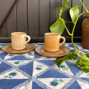 Op de afbeelding: Twee keramische koffiekopjes met schoteltjes. De kopjes zijn lichtbruin van kleur met een gespikkeld glazuur. De schoteltjes zijn donkerbruin van kleur met een gespikkeld glazuur. De kopjes en schoteltjes staan op een blauw-wit geblokt tafelkleed.