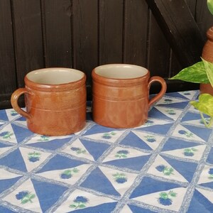 May include: Two brown ceramic mugs with white interiors and handles. The mugs are sitting on a blue and white checkered tablecloth with a floral pattern.
