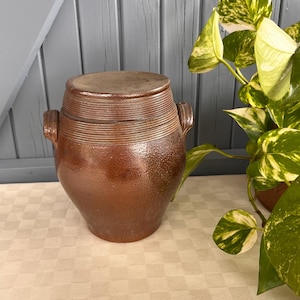 Pode incluir: Um pote de grés castanho com tampa, com detalhes horizontais canelados ao redor do corpo e pequenas alças. O pote antigo está sobre uma toalha de mesa xadrez, com uma planta verde e amarela ao fundo.