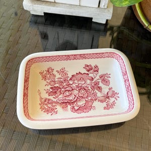Op de afbeelding: Een rechthoekige witte keramische schaal met een roze bloemenpatroon. De schaal heeft een verhoogde rand en een gegolfde rand. Het bloemenpatroon heeft roze bloemen, bladeren en een vogel.