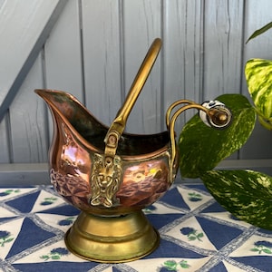 Può includere: Secchio per carbone antico in rame e ottone con una testa di leone decorativa e un manico. Il secchio presenta un motivo a diamante e una base in ottone. Il manico è in ottone con un dettaglio in ceramica bianca e blu. Su una tovaglia a fantasia blu e bianca.