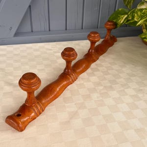 May include: A wooden coat rack with four rounded pegs. The rack is made of a warm-toned wood and is mounted on a wall. The pegs are evenly spaced along the length of the rack. A classic design for hanging coats, hats, or other items.