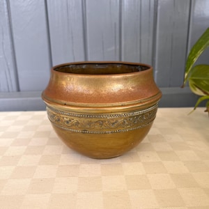 May include: A vintage copper bowl with a decorative band. The bowl has a rounded base and a flared rim. The band features an ornate, repeating pattern. The copper has a warm, aged patina, with variations in color from gold to brown.