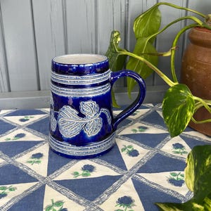 Op de afbeelding: Een kobaltblauwe keramische bierpul met een handvat, met witte blad- en wijnstokontwerpen. De pul heeft horizontale witte strepen en een getextureerde rand. Hij staat op een blauw-wit patroon tafelkleed, met een plant op de achtergrond.